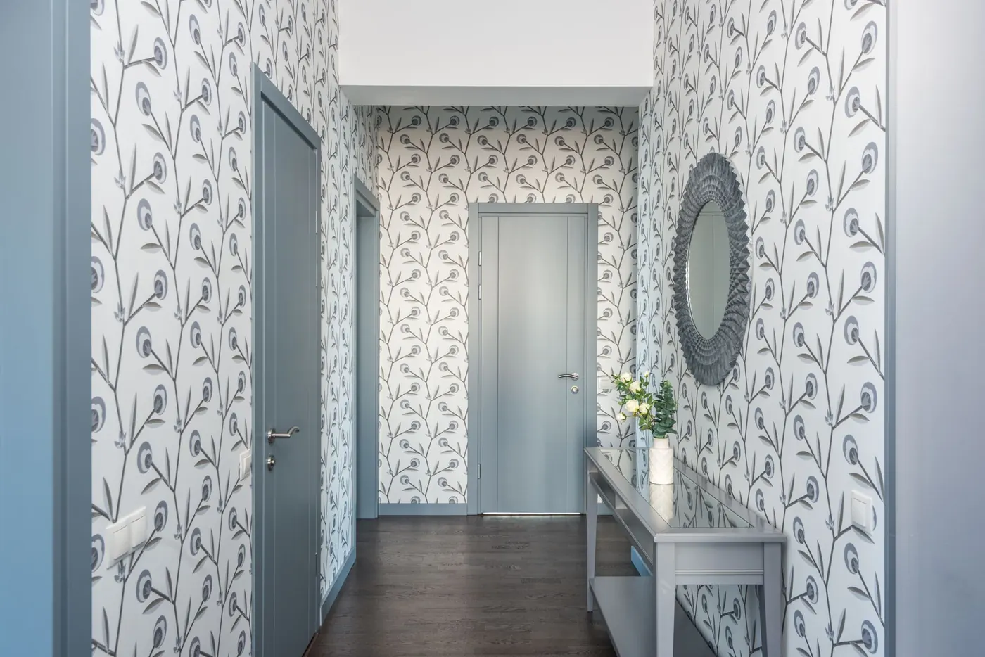 Bright hallway fully covered in a soft blue-grey botanical wallpaper, with a console table and round mirror